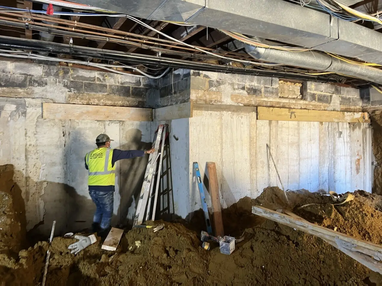 Construction worker in a safety vest standing near an step ladder inside an excavated basement area with concrete wall underpinning and exposed ceiling ductwork.