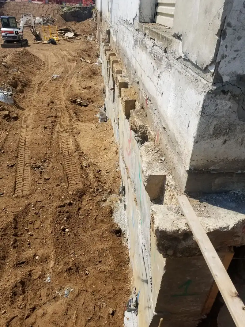 A high-angle, wide view looking down an outdoor excavation pit running alongside the concrete foundation of a large building undergoing concrete underpinning.