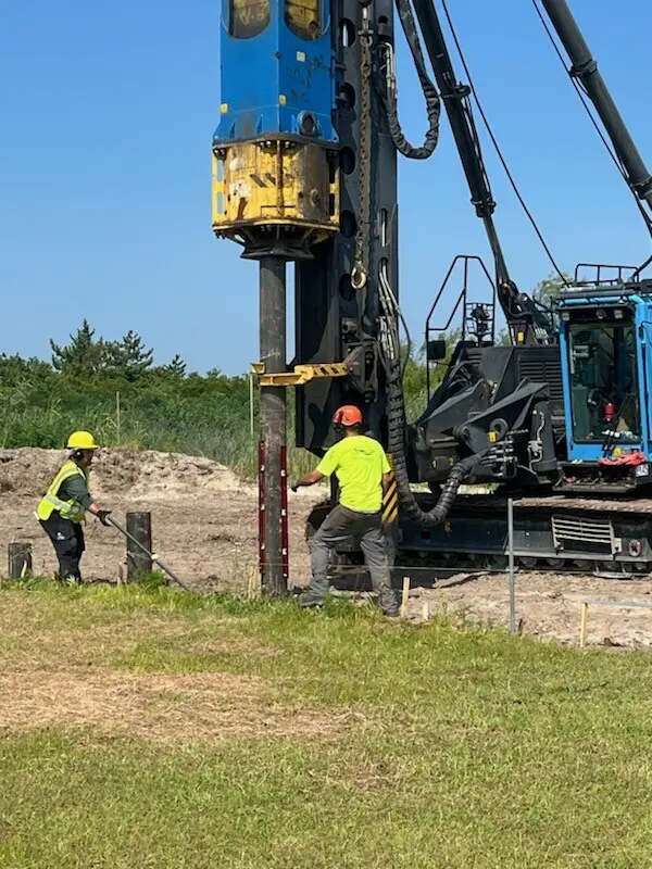 A close-up view of a large blue and yellow pile driving rig installing a steel foundation pile into the ground, while two construction workers wearing hard hats and high-visibility safety vests monitor and guide the operation on a grassy job site.