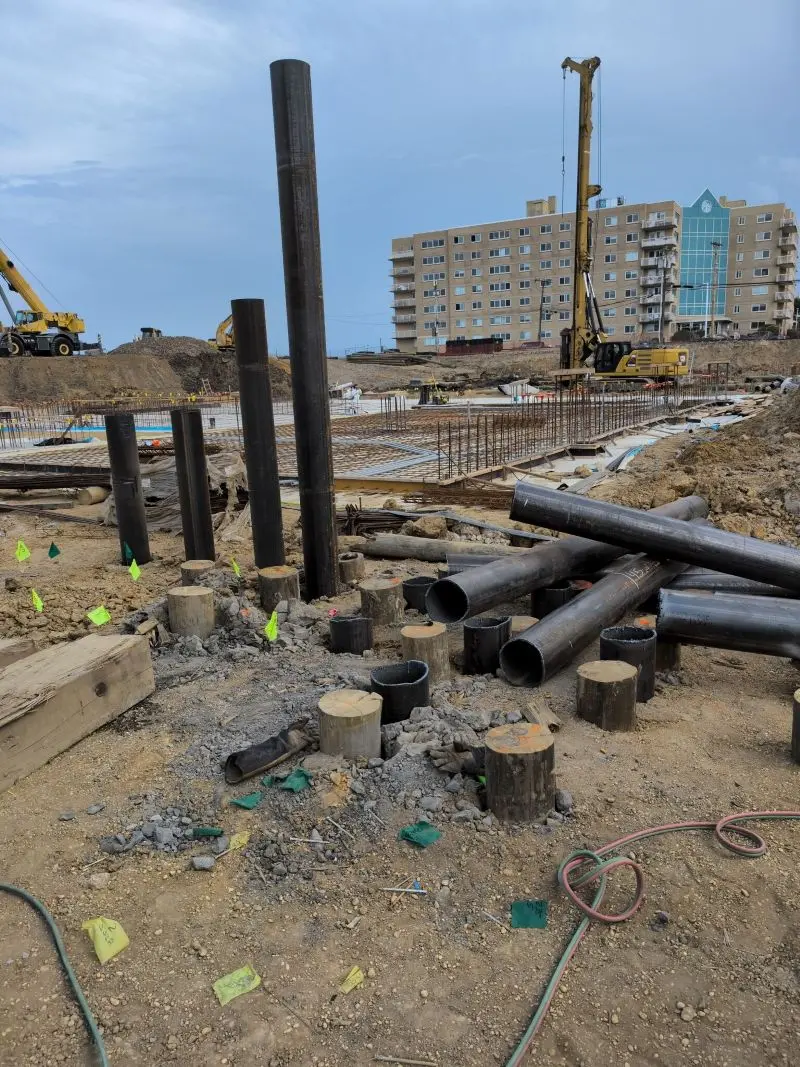 A busy commercial foundation construction site featuring several tall, vertical steel pipe piles and short wooden piles driven into the ground, with loose steel pipes scattered nearby, rebar grids laid out for a foundation, and heavy machinery, including an excavator and a crane, operating in front of a multi-story building under a cloudy sky.