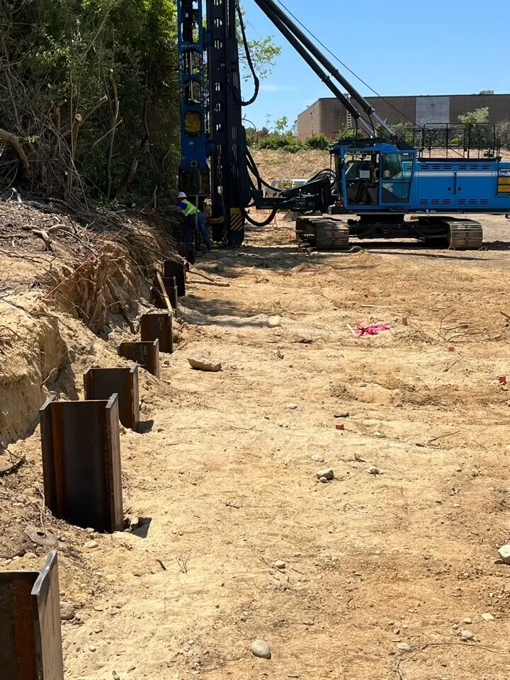 A row of heavy steel H-pile foundation beams driven vertically into the ground along a dirt embankment, with a large blue tracked pile driving rig operating in the background on a sunny day.