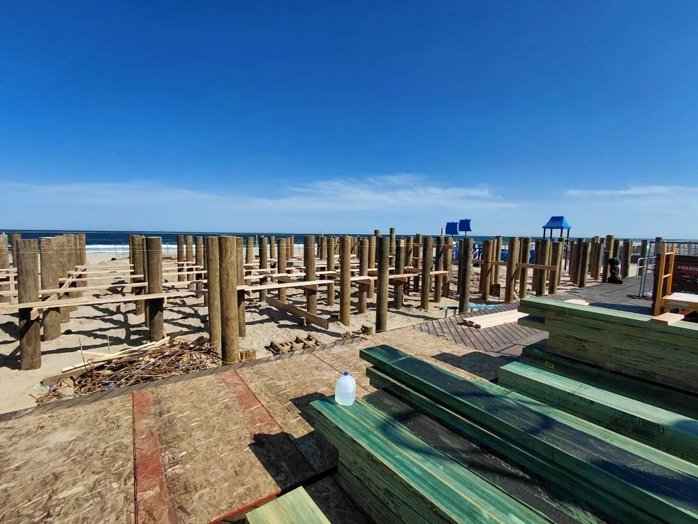 A wide construction site view on a sandy beach featuring a grid of thick wooden foundation pilings driven into the sand and connected by structural framing, with stacks of green treated lumber and plywood in the foreground under a clear blue sky.