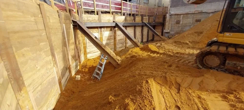Deep excavation site featuring timber lagging retaining walls supported by angled steel rake beams, with a tracked excavator standing on a large mound of sand.
