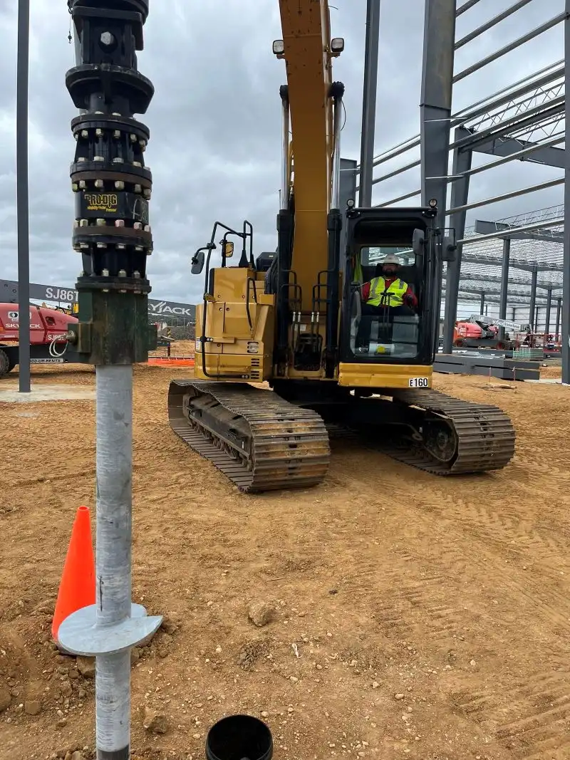 A large yellow tracked excavator with a hydraulic drive attachment installing a steel helical pile foundation into the ground at an active construction site, with an operator visible inside the cab and structural steel building frames in the background.