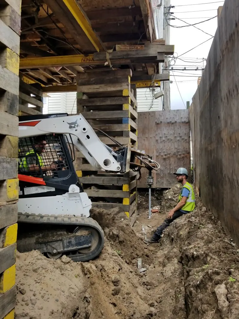A white tracked Bobcat skid steer loader equipped with a hydraulic drive attachment installs a steel helical pile into the ground under a raised building supported by wooden cribbing blocks, while a construction worker in a hard hat and high-visibility vest guides the process from a dirt trench.