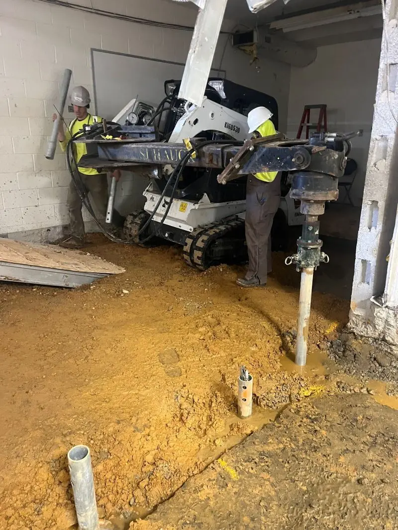 Construction workers operating a compact crawler drill rig to install helical pier foundation supports inside a dirt-floor building interior.