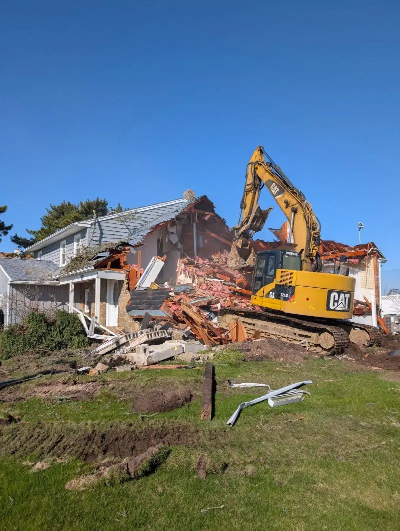 A large yellow CAT tracked excavator actively tearing down a two-story residential house on a demolition site, with the structure partially collapsed into a pile of wooden debris and rubble under a clear blue sky.