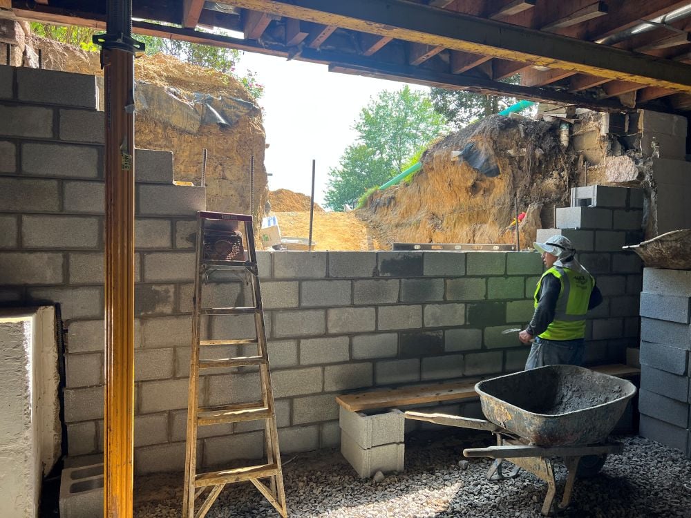 An indoor, eye-level view from underneath an elevated structure or basement undergoing foundation reconstruction, focusing on a mason building a new concrete block wall. On the right, a construction worker wearing a high-visibility safety vest and a sun hat stands next to a metal wheelbarrow filled with wet mortar, working on the partially completed concrete masonry unit (CMU) wall.