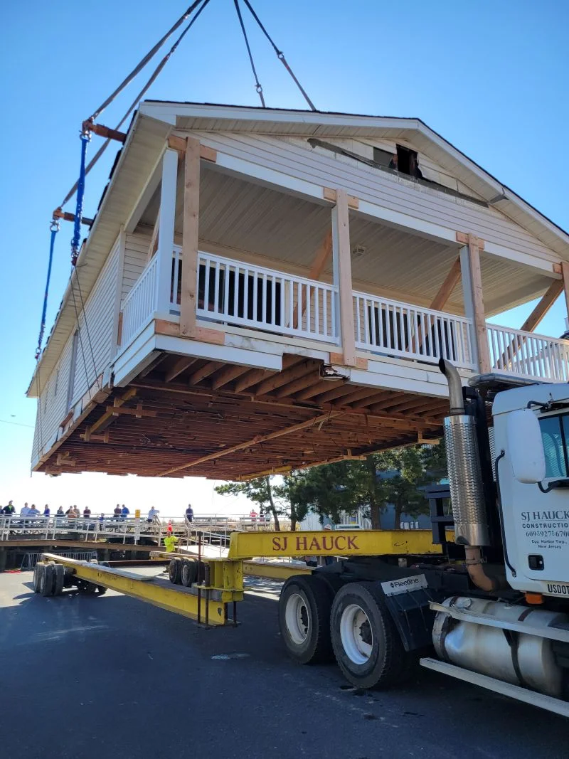 A entire two-story house with light siding and a covered front porch suspended mid-air by a crane system, positioned directly over a long yellow flatbed truck trailer labeled "SJ HAUCK".