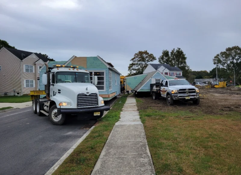 Heavy-duty white transport trucks moving sections of a light-blue modular home down a residential street.