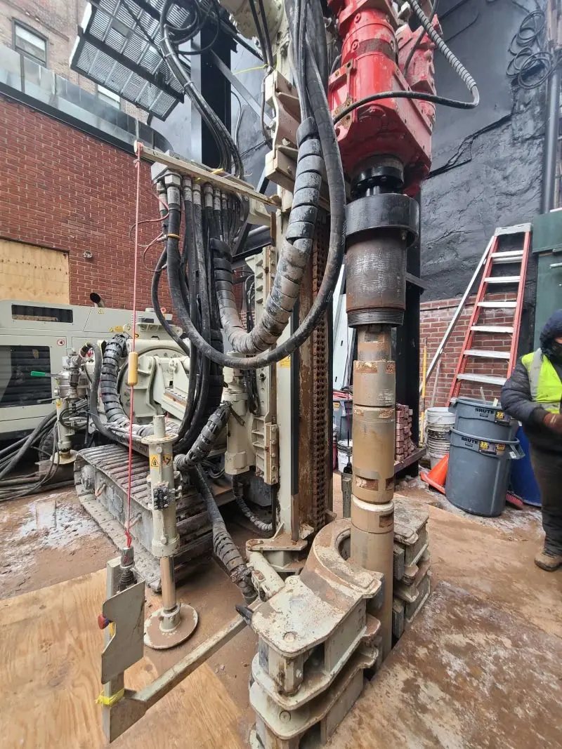 A high-angle view of a compact tracked drilling rig utilizing a heavy hydraulic rotary head to install steel casing segments for micropiles into the ground within a tight, confined outdoor workspace between brick buildings.