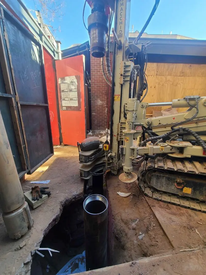 A high-angle view of a compact tan tracked drilling rig positioned next to a narrow trench, with a heavy hydraulic rotary head aligned above a threaded steel casing segment being drilled into the ground for a micropile foundation at a tight urban construction site.