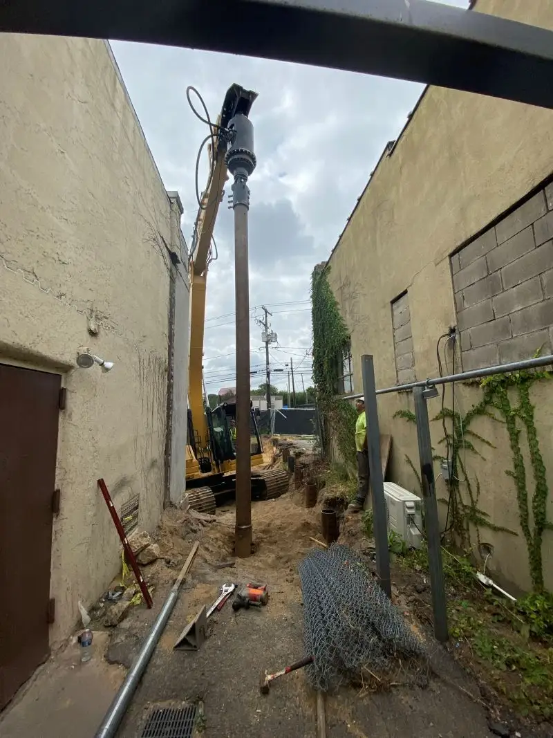 A yellow CZM 317 tracked drilling rig drilling deep into the ground on an elevated construction site next to a concrete wall and residential fence.