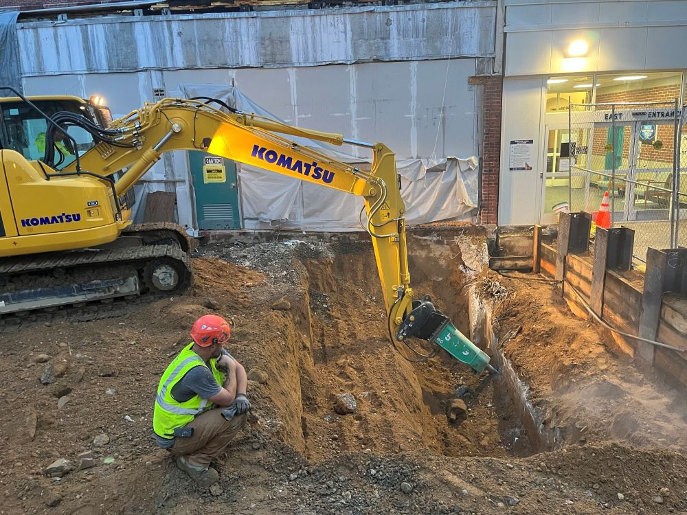 Komatsu excavator with a hydraulic breaker attachment digging a trench while a worker observes.