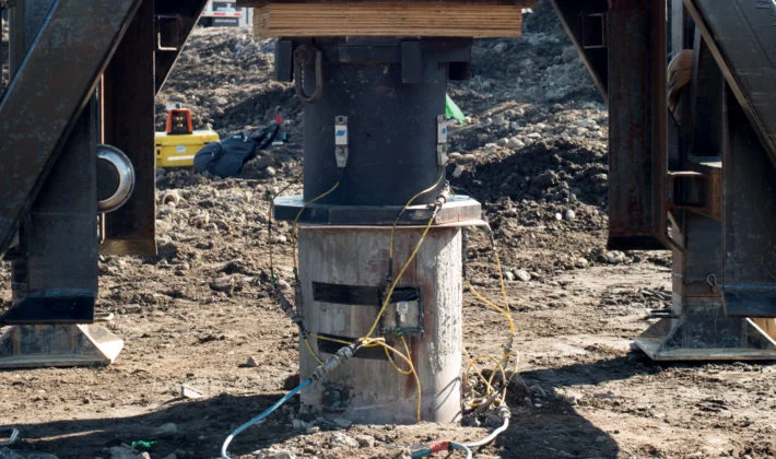 Close-up view of a heavy hydraulic jack and sensor testing apparatus mounted on top of a concrete foundation pile at a construction site.