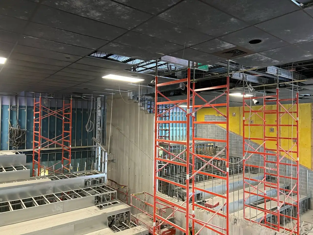 Interior renovation site featuring bright orange metal scaffolding towers reaching up to a dark drop ceiling inside a multi-level commercial space with exposed metal framing.
