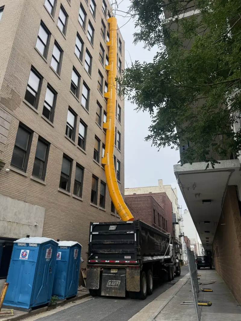 Tall yellow construction trash chute attached to the side of a multi-story brick building, feeding directly into a large black dump truck parked below.