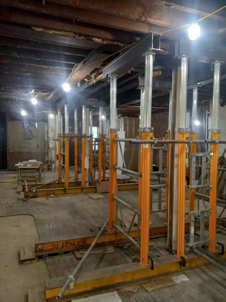An indoor, medium-angle view of a construction or renovation site inside an older building, featuring multiple heavy-duty structural shoring towers. The shoring systems consist of bright orange vertical main posts topped with adjustable silver screw jacks, which extend upwards to support heavy horizontal steel I-beams running directly beneath the exposed wooden ceiling joists. The system is braced together with silver metallic scaffolding tubes and couplers, and the entire assembly rests on thick steel or wooden sill beams on the floor to distribute the massive load. Temporary string lights illuminate the dim, open workspace.