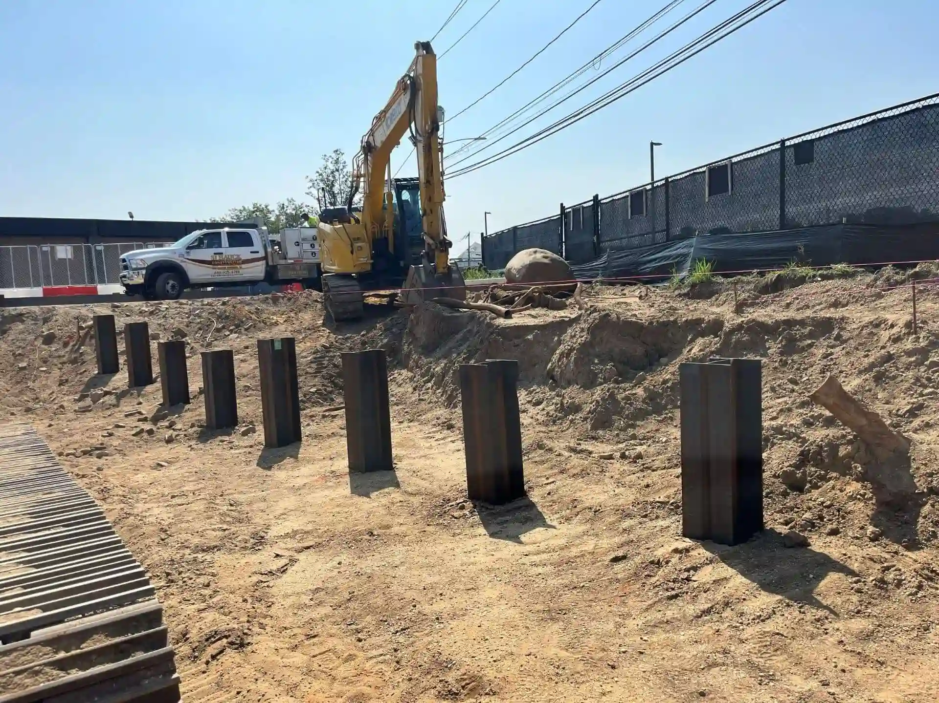 A low-angle view of a construction site featuring a row of heavy steel H-pile foundation beams driven vertically into the dirt ground, with a yellow tracked excavator and a white work truck operating on an embankment in the background under a clear sky.