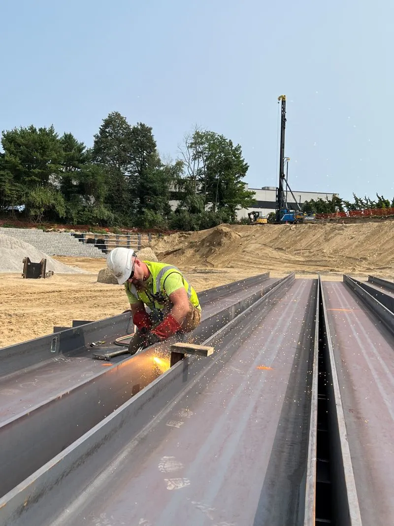 Construction worker in a high-visibility vest and hard hat using a cutting torch on large steel I-beams at an outdoor excavation site.