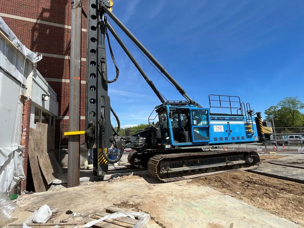 Large blue tracked foundation drilling rig operating adjacent to a multi-story brick building.
