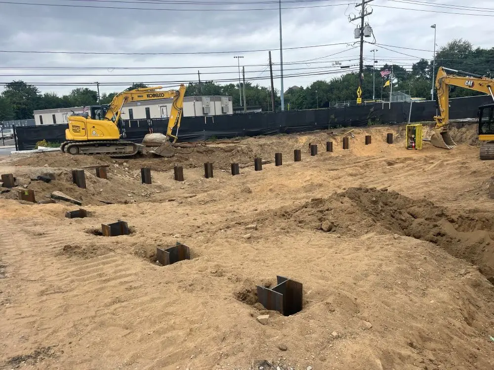 A wide view of an excavated construction site featuring rows of heavy steel H-pile foundation beams driven vertically into the dirt ground, with two yellow tracked excavators operating on the dirt terrain under an overcast sky.