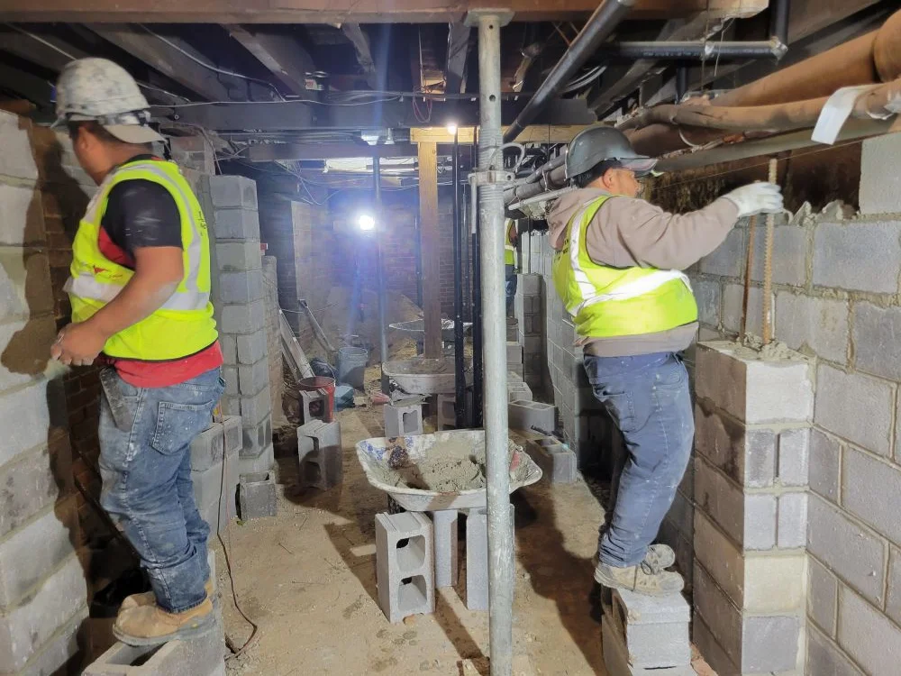 A construction worker wearing a high-visibility vest and hard hat walking up wooden steps from a basement work area, beneath a heavy yellow steel beam labeled "S.J. HAUCK" supported by adjustable metal shore posts.