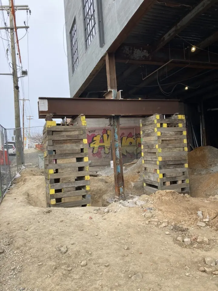 Heavy brown steel I-beam supported by two tall wooden cribbing stacks, providing temporary structural support under the corner of a building undergoing excavation work.