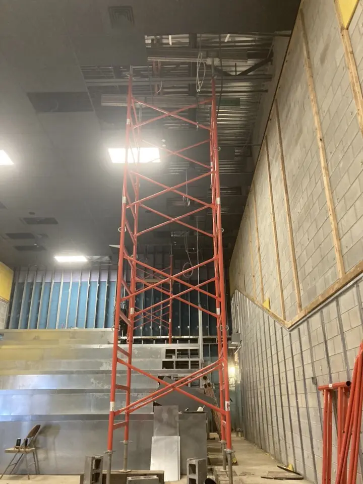 A tall, bright orange metal scaffolding tower reaching up into an open section of a dark drop ceiling inside a commercial building undergoing interior renovation, situated next to a high concrete block wall and tiered structural framing.