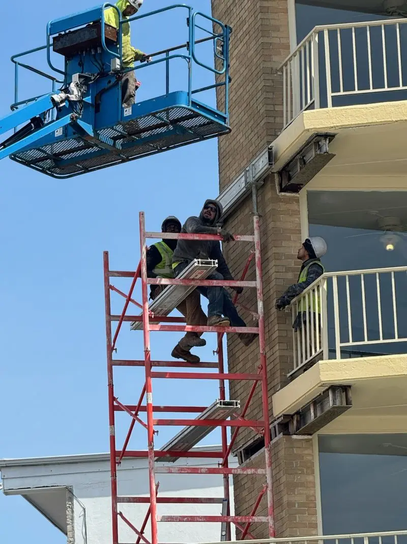 Construction workers operating on a red metal scaffolding frame and in a blue boom lift basket alongside the exterior balcony of a multi-story brick building.