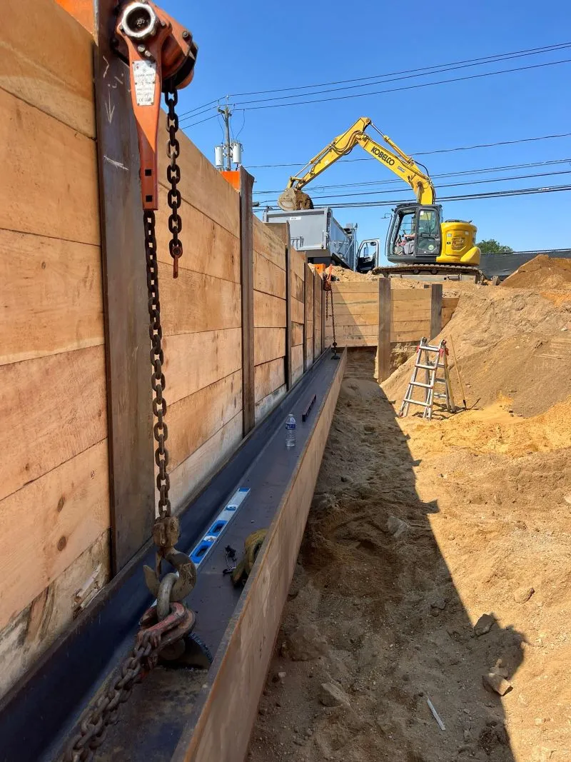 Deep trench excavation site showing timber lagging retaining walls, steel beams with chain hoist equipment, and a yellow Kobelco excavator working in the background.