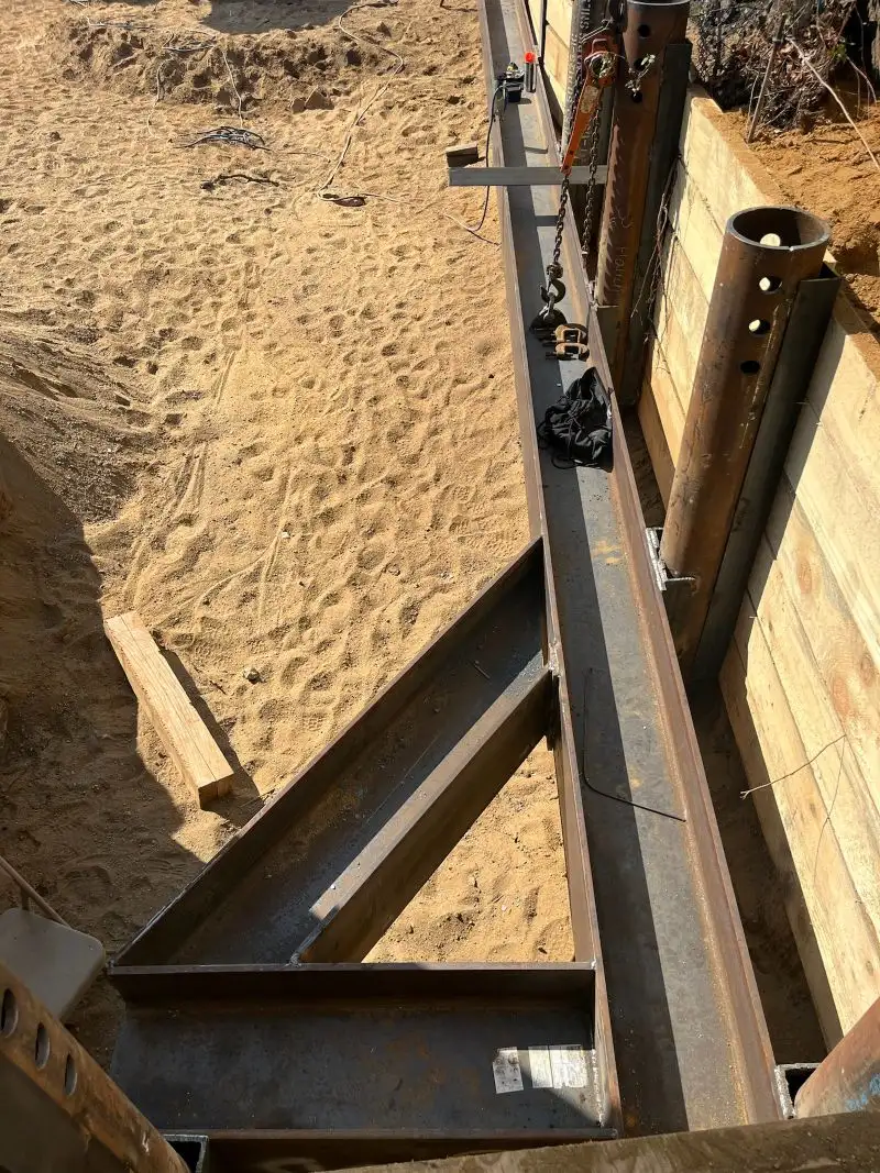Overhead view of a steel shoring and bracing framework installed along a sandy excavation pit next to a wooden retaining wall.
