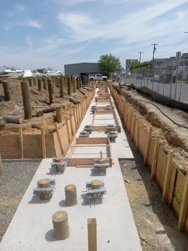 A high-angle view looking down a long, narrow excavated trench lined with wooden shoring walls. A fresh concrete footing slab runs along the bottom of the trench, with short wooden timber piles protruding up through it. Many of the piles are fitted with heavy steel bracket assemblies or caps, and wooden formwork boxes are positioned at intervals along the concrete path. On the higher dirt bank to the left, rows of taller timber piles stand driven into the ground, with boats and marina buildings visible in the background under a partly cloudy sky.