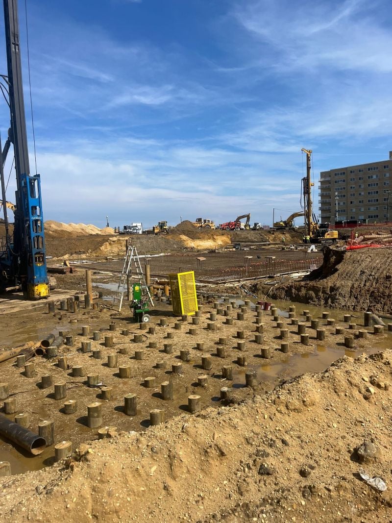 A wide, high-angle view of a large commercial construction site featuring a dense grid of short wooden timber piles driven into muddy ground in the foreground, with a large blue pile driver on the left, an excavated foundation area with rebar in the center, and several yellow excavators operating near a multi-story building under a blue sky with light clouds.
