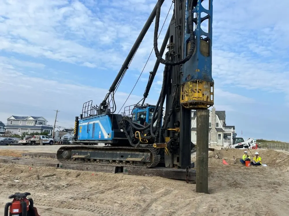A large blue and black tracked pile driving rig positioned on a sandy construction site, actively driving a thick wooden timber pile vertically into the ground. In the background, two workers in high-visibility vests sit on a dirt embankment near residential houses under a partly cloudy sky.